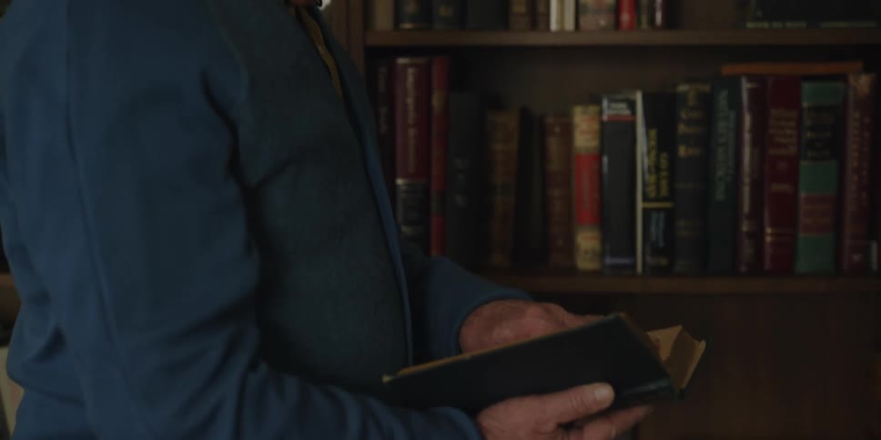 A man takes a book from his shelf and opens and it thinking as he walks away from the bookcase