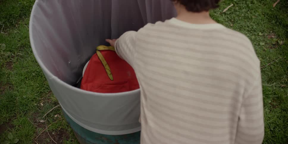 A boy stands over an outdoor garbage can and takes out a backpack and picks up a drawing from the ground