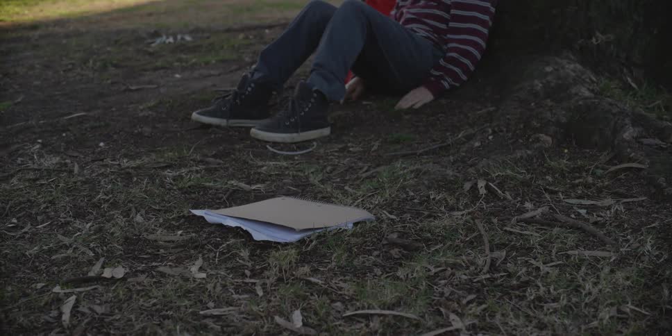 A young boy leaning against a tree outside throws a stack of papers on the ground