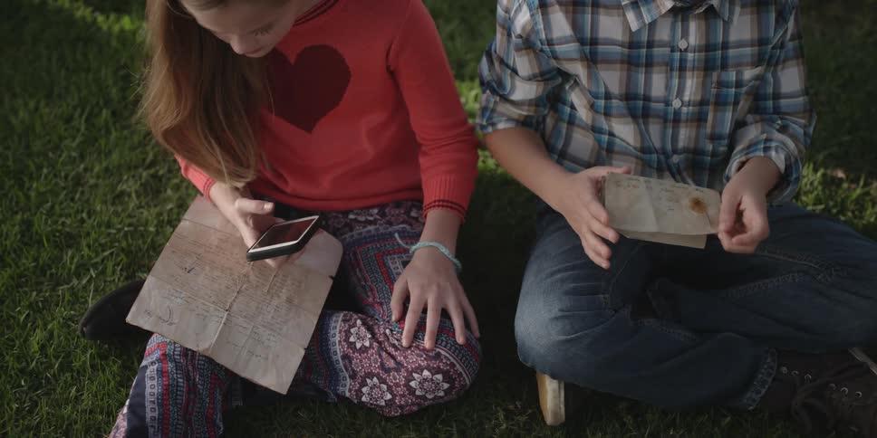 A young boy sits on the grass outside and picks a handwritten note out of a box and opens it while the girl sitting next to him holding a note looks at her cell phone