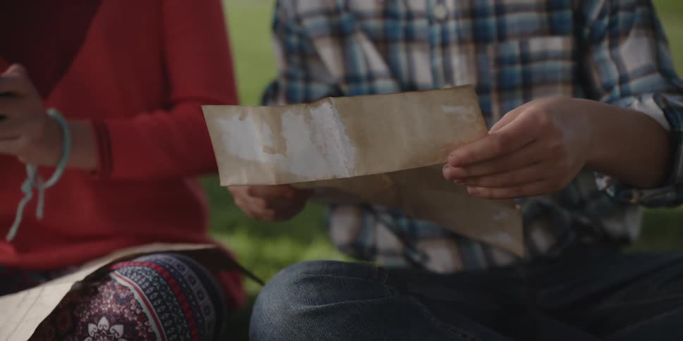 Two teenagers are sitting outside on the grass and he is holding a paper and she is holding her phone