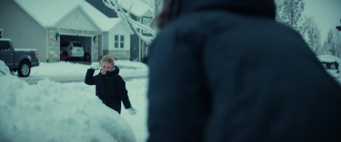 A man is outside in his driveway as his young daughter throws a snowball at him