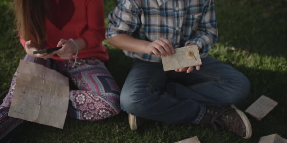 A girl and boy are sitting and reading papers while the girl uses her phone