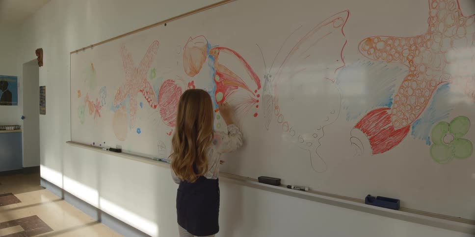 Girl with wavy brown hair white shirt and black skirt draws butterfly on whiteboard in the classroom