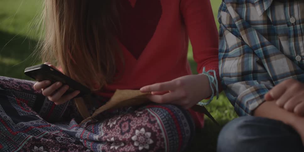 Woman wearing red shirt and pants sits on green lawn next to man in plaid shirt and jeans reading a sheet of paper