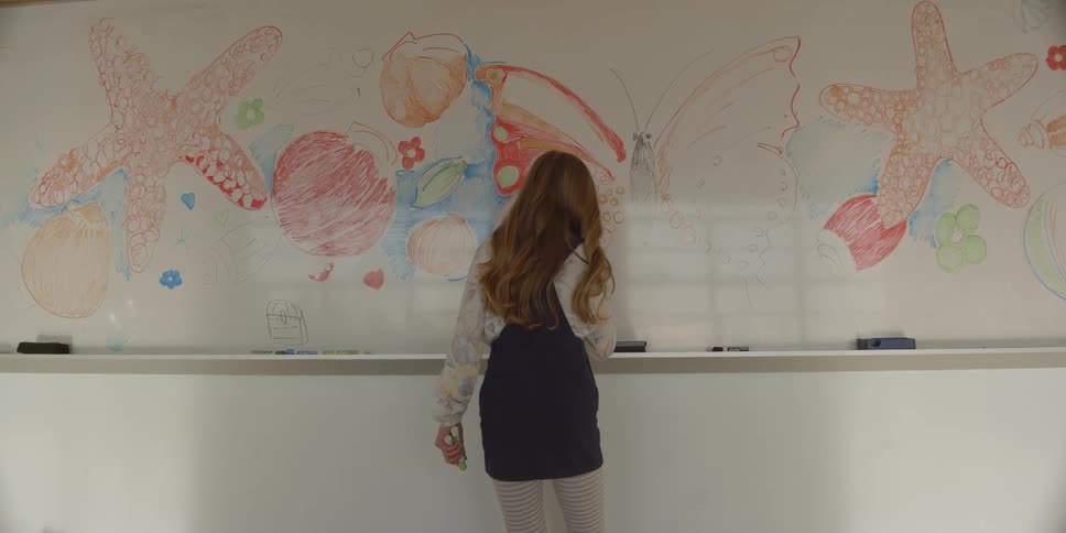 Rear of girl wearing striped leggings and blue skirt draws on whiteboard in the classroom
