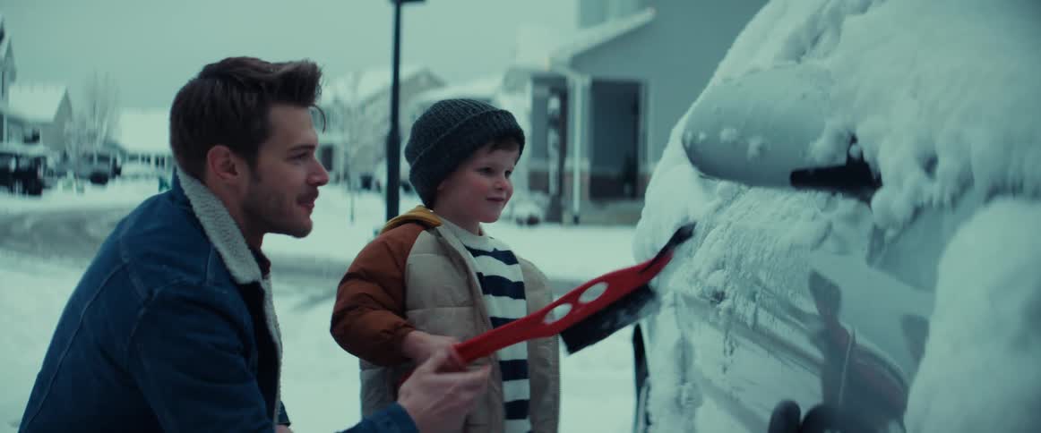 A man watches as a child attempts to clean snow off of a car with a snow brush