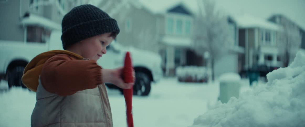 A father walks past with a shovel as his son sweeps snow off the car