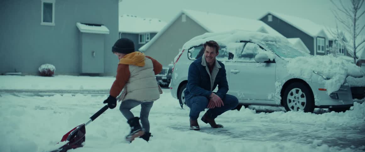 A dad kneels down in the driveway and smiles and points as his young son shovels snow