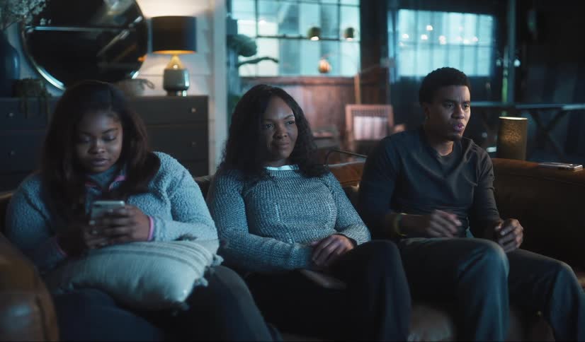 A family viewing their smartphones while sitting together on a sofa look up one by one to focus on the television