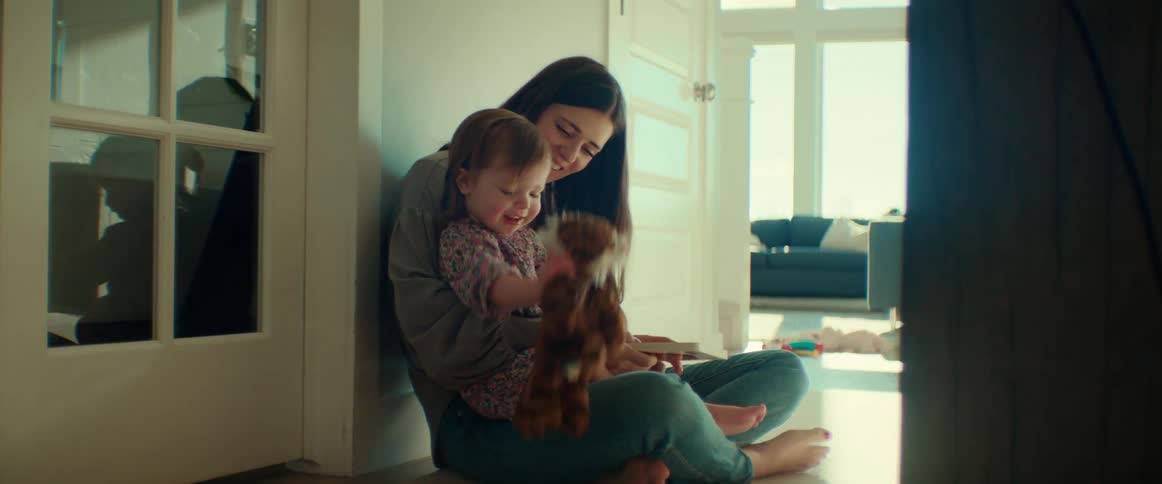 A young woman sits on the floor while holding a young girl in her lap as the girl plays with a plush animal
