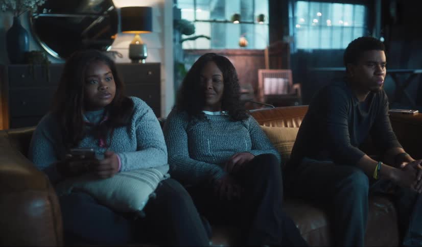 A family are sitting on the couch in front of the television looking at their phones and they all put the phones down at the same time and look at the television