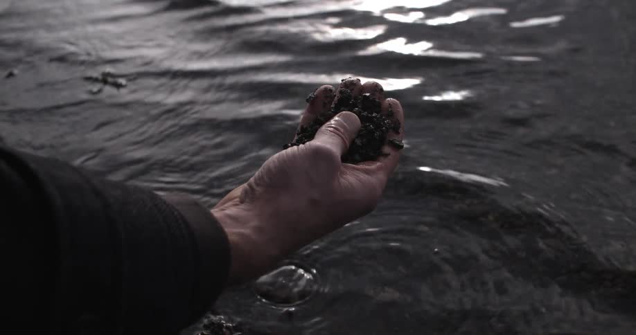 A man grabs a fistful of rocks and mud from a shoreline
