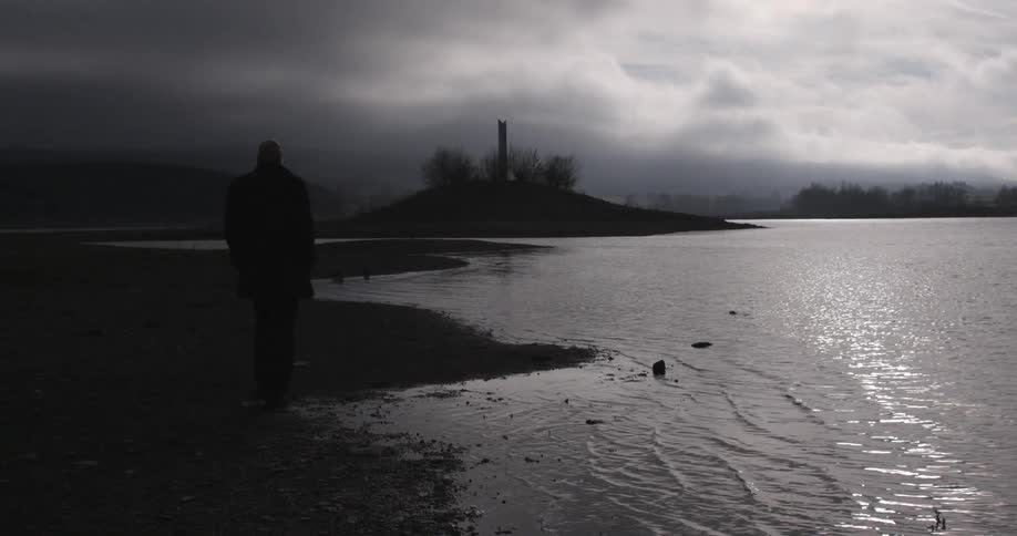 A man walking along a rocky shoreline as night falls under a stormy sky
