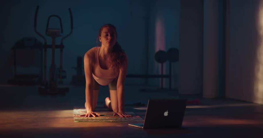 Woman with ponytail sport shorts and top stretches on yoga mat watching laptop in gym