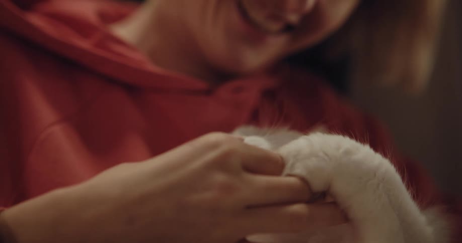A woman is holding her white cat and feeding it a treat as it puts its paw on her hand