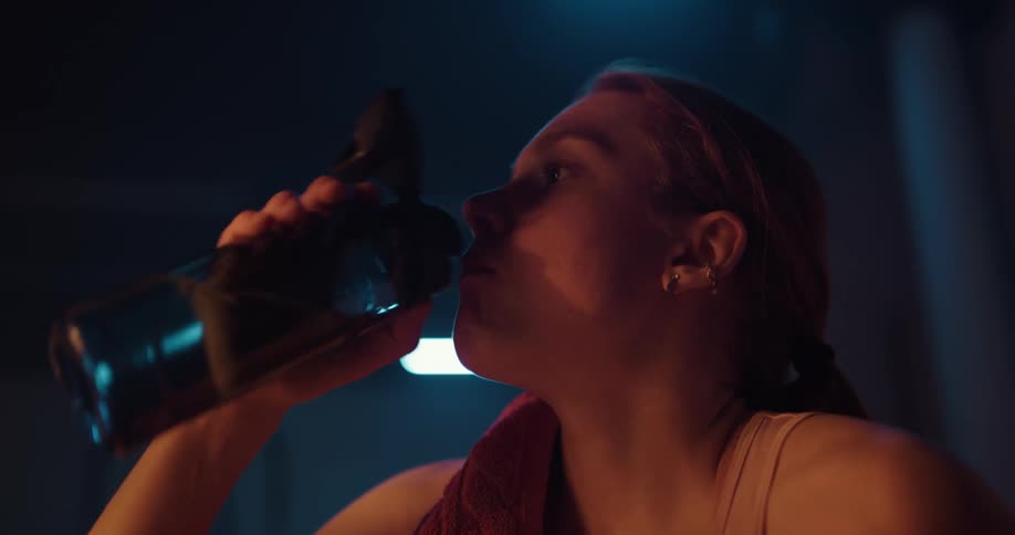 Blonde woman with white top and towel on shoulder drinks water from bottle after working out