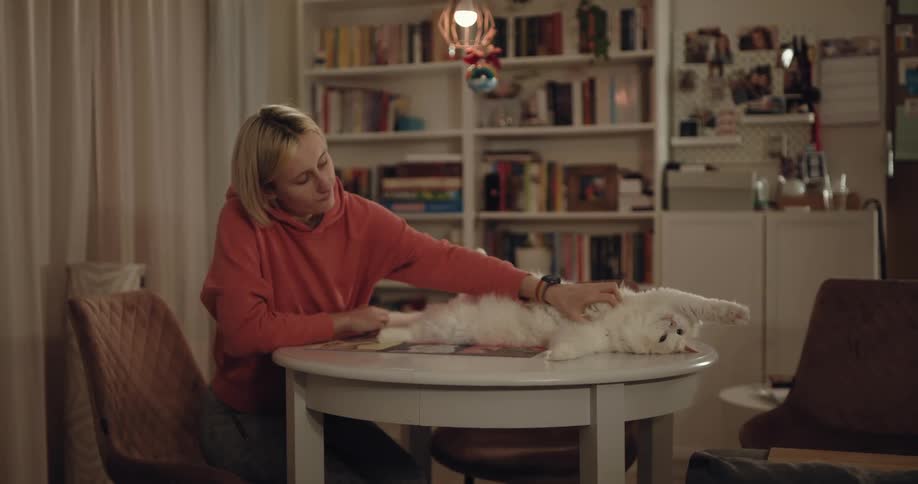 Blonde woman with bob and orange hoodie pets white cat lying down on white wooden table in room
