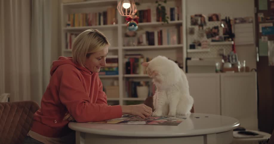 A womans cat sits on the table while she works on a jigsaw puzzle