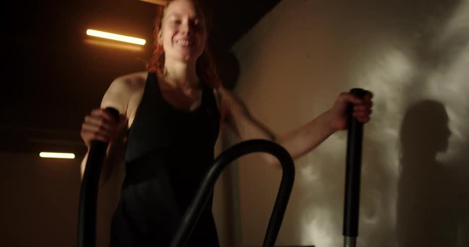 Woman with ponytail black top and shorts uses cross trainer and smiles at camera in gym