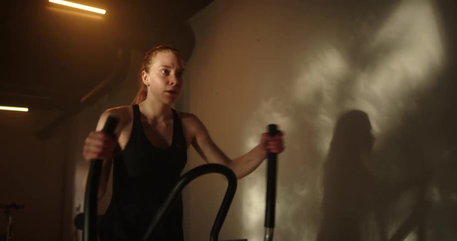 Determined woman with ponytail black top and shorts uses cross trainer at gym
