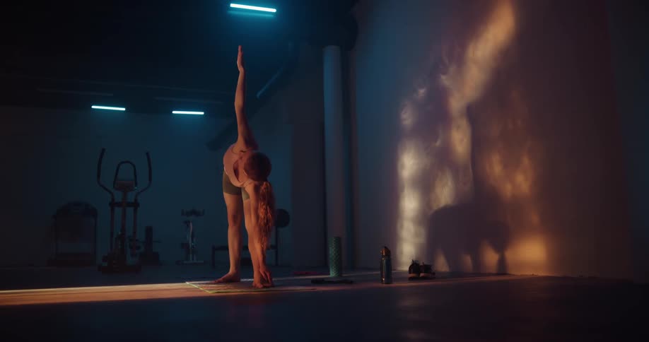 A woman is standing on a mat in a room stretching her arms out and touching her toes