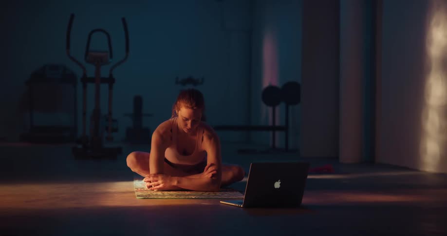 A woman is sitting on the floor with her legs crossed and doing arm movements in front of a laptop computer on the floor
