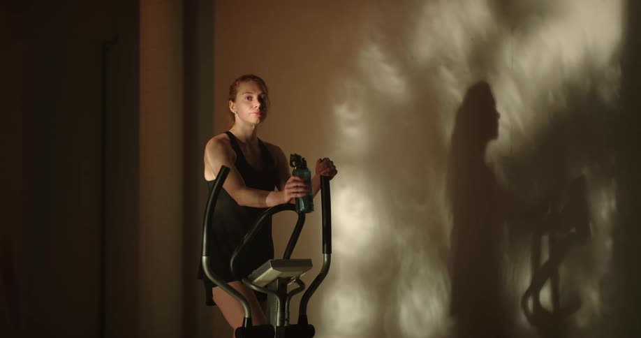 A woman drinks water while sitting on an elliptical machine