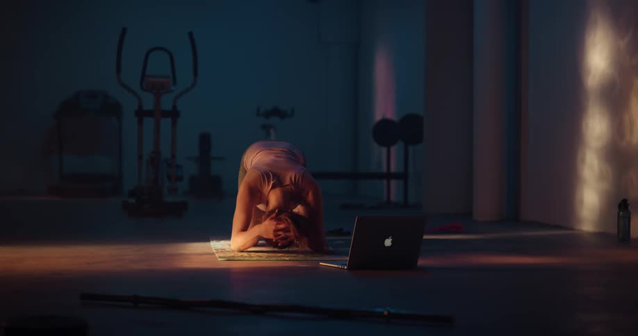 Woman with shorts and pink top watches laptop and does stretching at gym