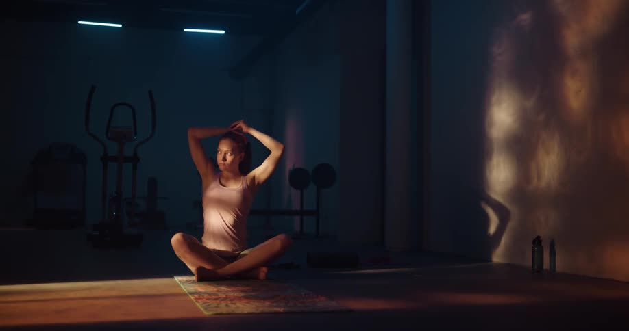 A woman is sitting on the floor on a mat with her legs crossed as she shakes her head and pulls back her hair
