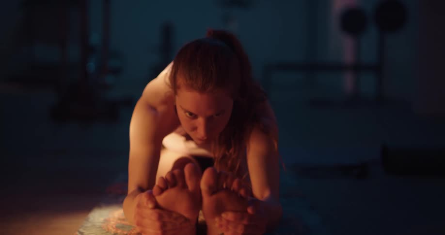 Red haired woman clasps her feet while bending for yoga stretch on yoga mat in dim home gym