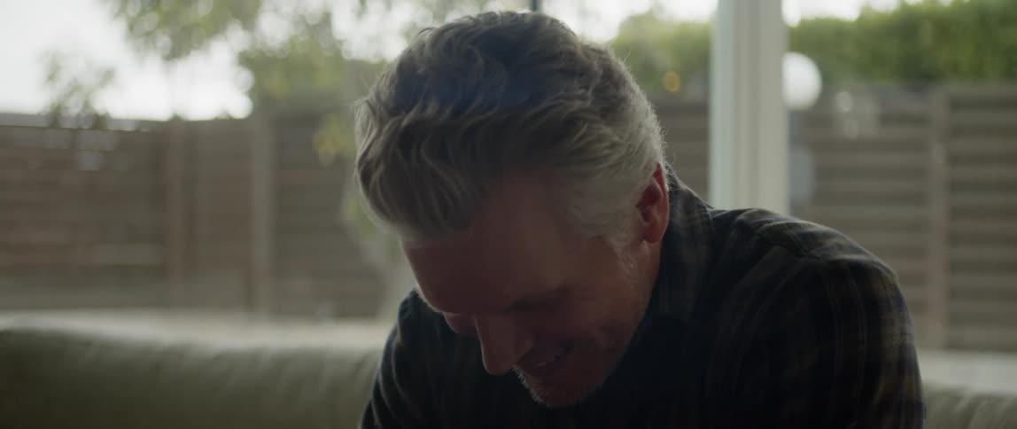 Middle aged man in dark green plaid shirt sits on couch talking and smiling in living room