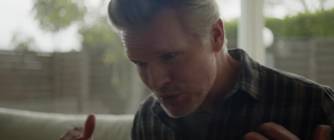 A man is sitting inside on a couch near windows smiling and moving his arms as he talks
