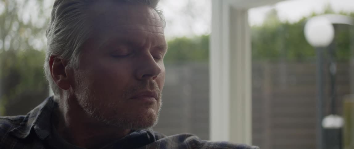 Middle aged man with beard and gray hair and dark green plaid shirt sits on couch closing his eyes and thinking in the living room at home