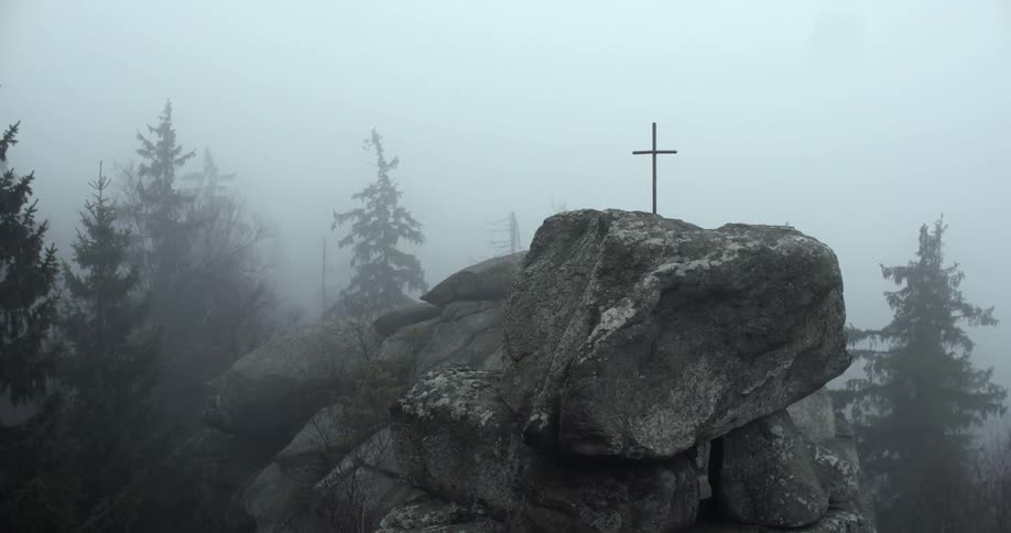 Fog creeps around a rock formation with a cross on top