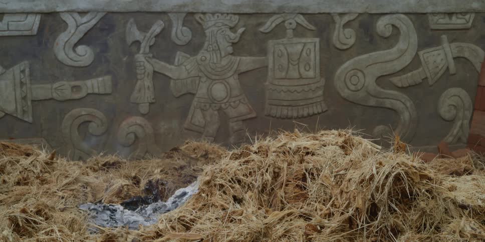Stack of burning dry hay or grass smoldering in front of a wall carved with aztec symbols