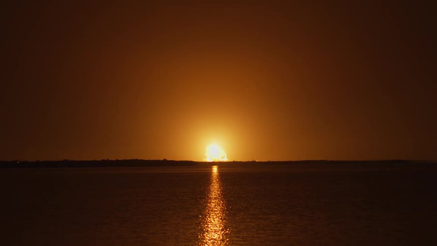 A rocket takes off in the darkness as a glowing light rises on the horizon and leaves a plume of smoke behind it as the light reflects on the ripples in the water