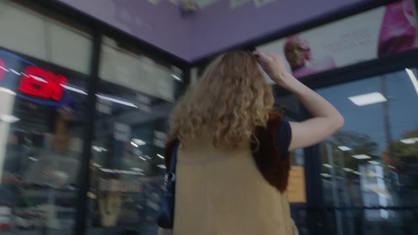 A woman is walking on a city sidewalk and turns to go into a store and pulls her sunglasses on top of her head as she walks in the door