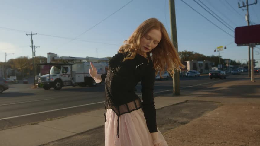 Woman with red hair black shirt and white skirt dances in the street near highway