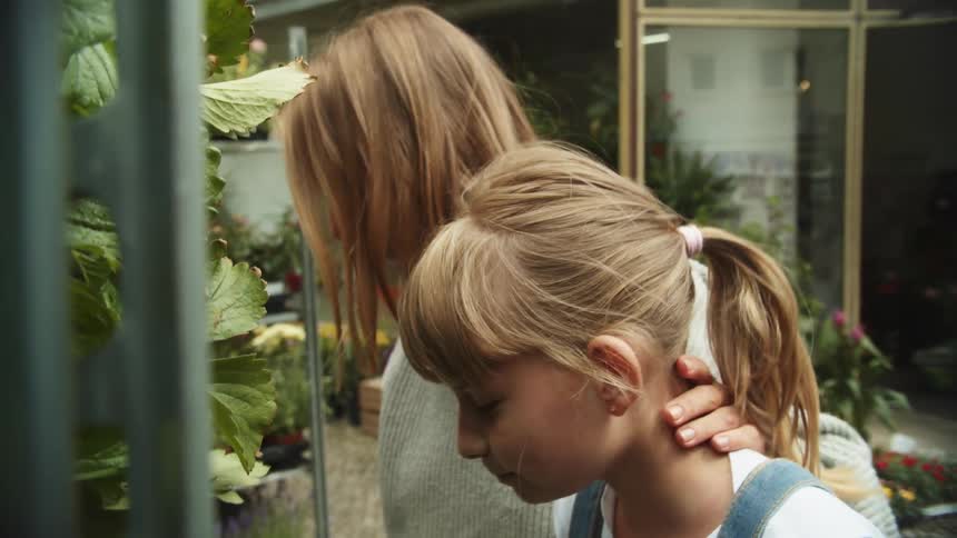 A woman and a girl standing by shelves with plants with the girl eating a fruit