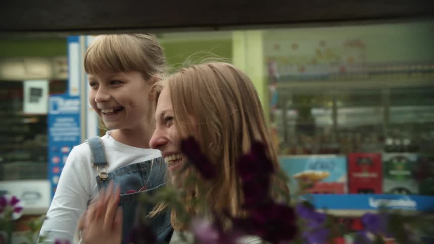 A woman picking up a girl in a store with flowers in the foreground and shelves in the background