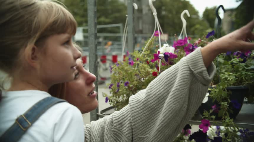 Mother carrying her daughter on her arms looks at flowers on a stand in the streets of a small town