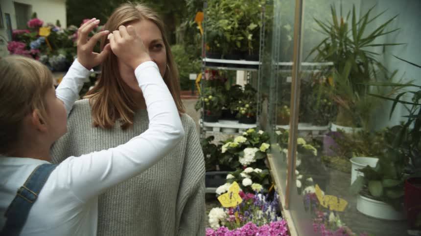A young girl places a small flower in a womans hair while they stand outside in a small garden