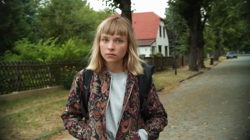 A woman with a jacket and a backpack walking in a neighborhood with houses trees the sky and clouds in the background