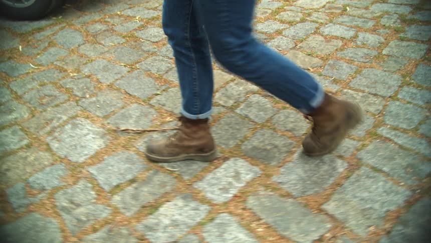 A pair of a womans legs wearing blue jeans and brown boots walking outdoors at daytime on cobblestone pavement