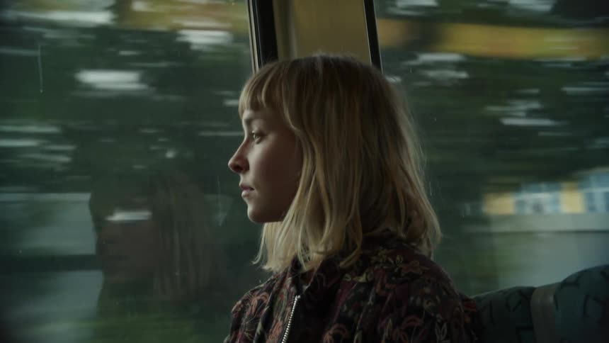 A woman with a jacket sitting on a train with buildings the sky and clouds in the background