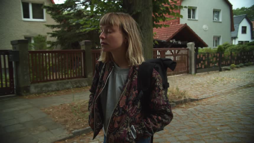 A woman with a jacket and a backpack walking in a neighborhood with trees and houses