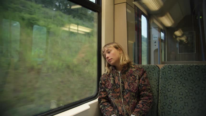 A young woman sitting on a moving train laying her head against the window staring outside