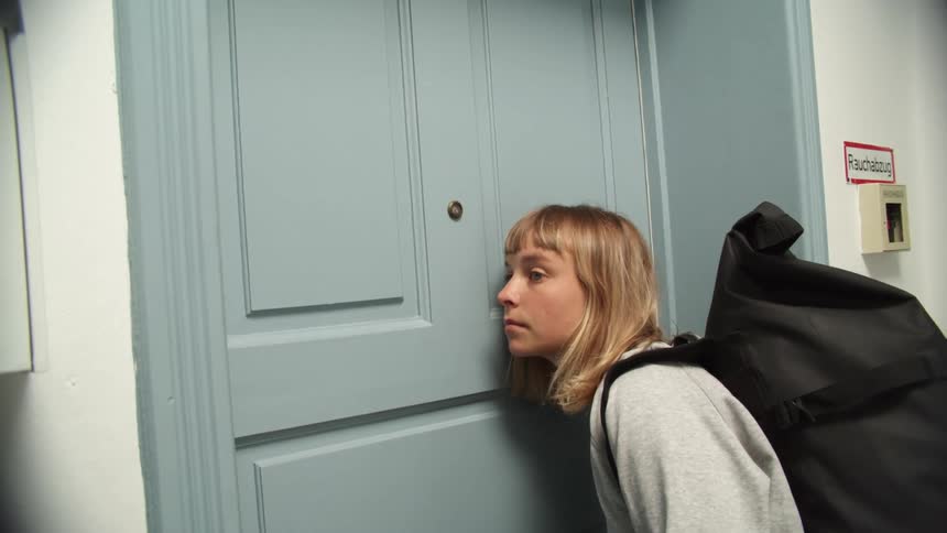 A teenage girl putting her ear up to a blue apartment door while wearing a backpack