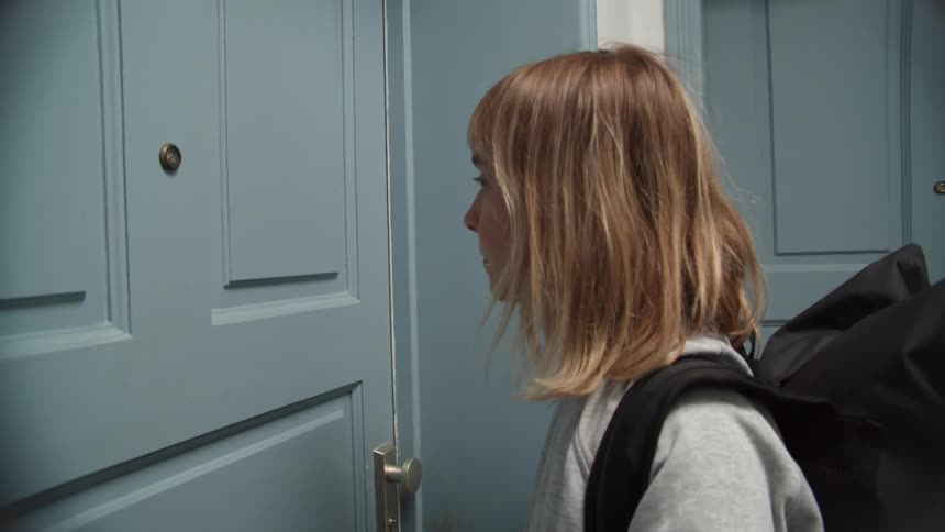 A woman with a sweater and a backpack in a hallway knocking on a door
